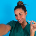 Beautiful Doctor Woman Wearing Medical Uniform Over Blue Background Make Selfie Holding An Invisible Braces Aligner, Recommending This New Treatment. Dental Healthcare Concept.