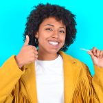 Young Woman With Afro Hairstyle Wearing Yellow Fringe Jacket Over Blue Background Holding An Invisible Braces Aligner And Rising Thumb Up, Recommending This New Treatment. Dental Healthcare Concept.