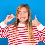 Caucasian Teen Girl Wearing Striped Shirt Over Blue Studio Background Holding An Invisible Braces Aligner And Rising Thumb Up, Recommending This New Treatment. Dental Healthcare Concept.