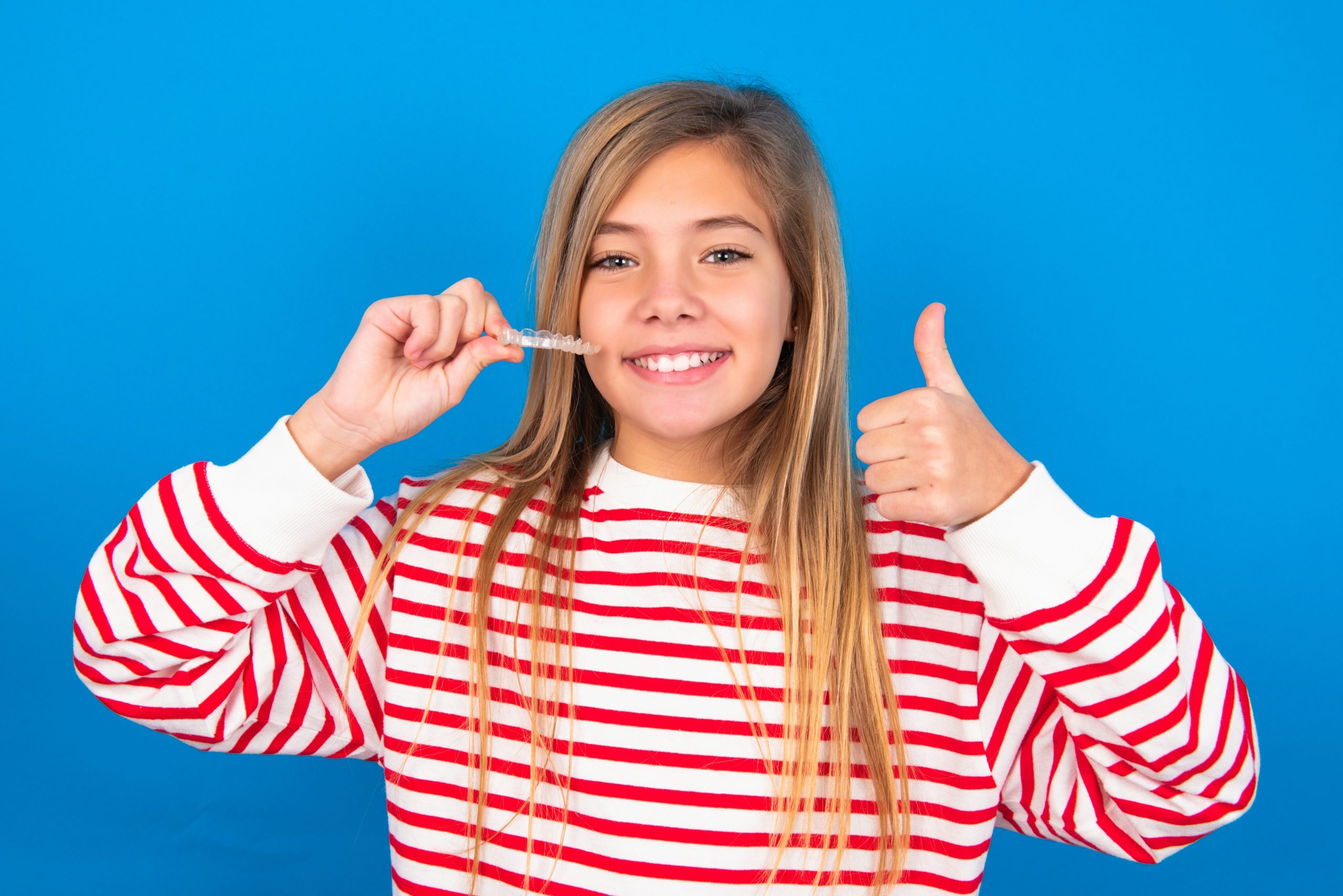 Caucasian Teen Girl Wearing Striped Shirt Over Blue Studio Background Holding An Invisible Braces Aligner And Rising Thumb Up, Recommending This New Treatment. Dental Healthcare Concept.